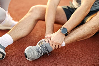 Athlete sitting on a running track, holding his injured ankle, feeling intense pain after a sports accident, and now seeking care from a Henry Ford Sports Medicine specialist. 