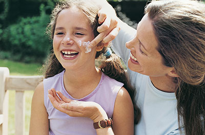 mom smiling and applying sunscreen to her young daughters cheek as they both are laughing