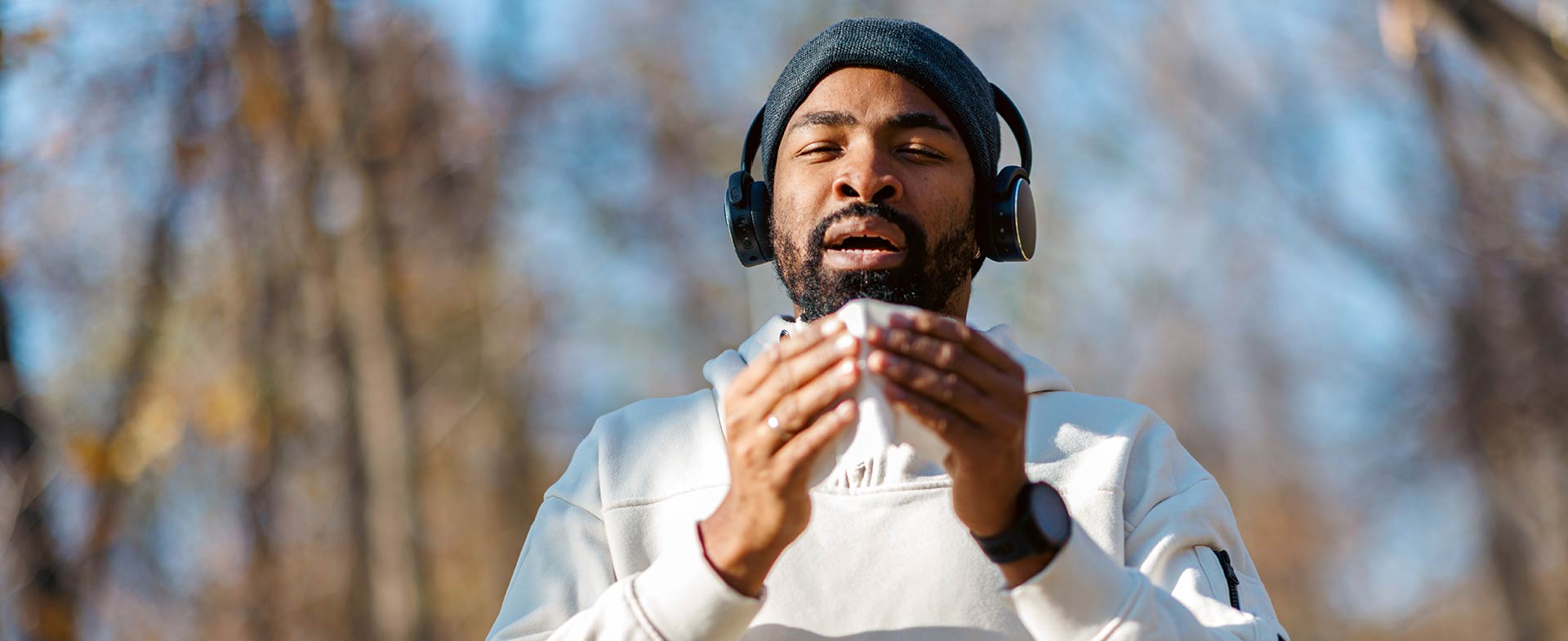A man standing outdoors in a forested area holding a tissue, ready to sneeze. They are wearing headphones, a light-colored hoodie and a beanie. The background shows bare trees and a clear blue sky, suggesting a cool day. working out sick