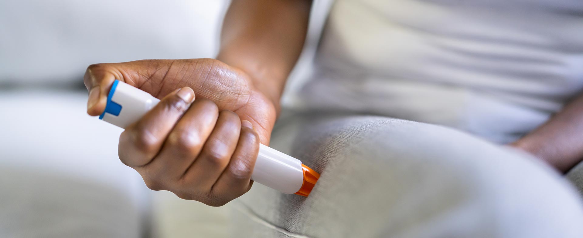 A person seated on a couch is using an epinephrine auto-injector (EpiPen) on their thigh, depicted in a close-up shot. The individual wears a light-colored shirt and pants. epipen interactions