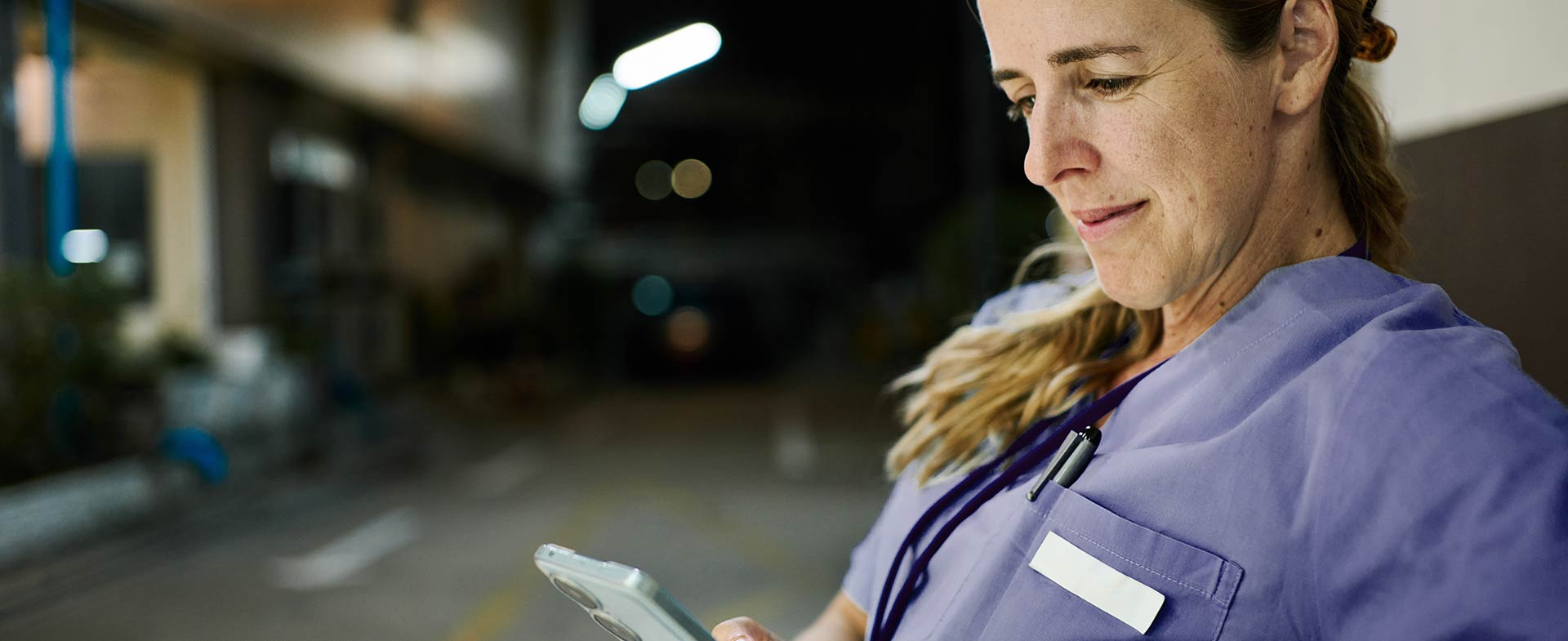 A woman with a blonde ponytail in medical scrubs is seated outdoors, looking at a smartphone. The background suggests a hospital environment at night. digital interventions for night shift workers