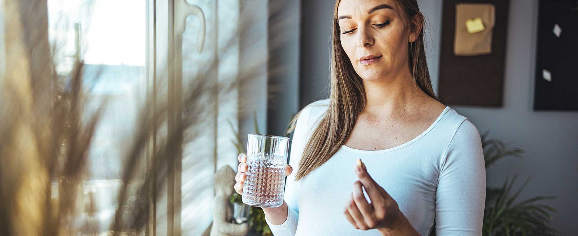 A pregnant woman with long hair and a long sleeve white maternity shirt holds a glass of water and a white pill. She stands inside near a window with bright natural light. aspirin use in pregnancy