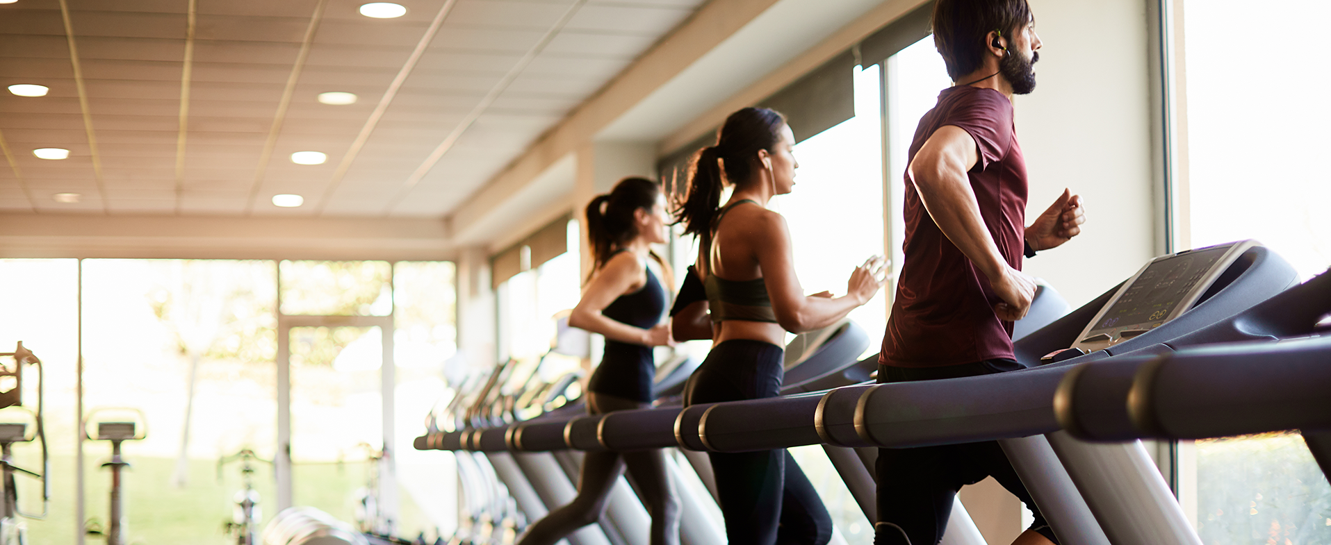 Two women and one male at a gym running on treadmills. They are looking out the window and wearing workout clothes. people running on treadmill