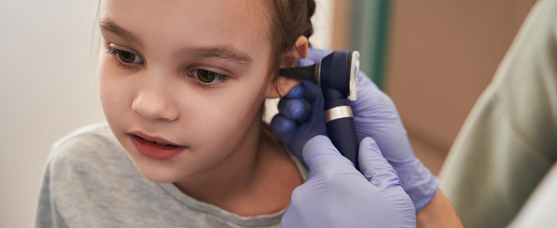 A person wearing blue gloves examines a young girl's ear with an otoscope in a medical setting. kid ear infections