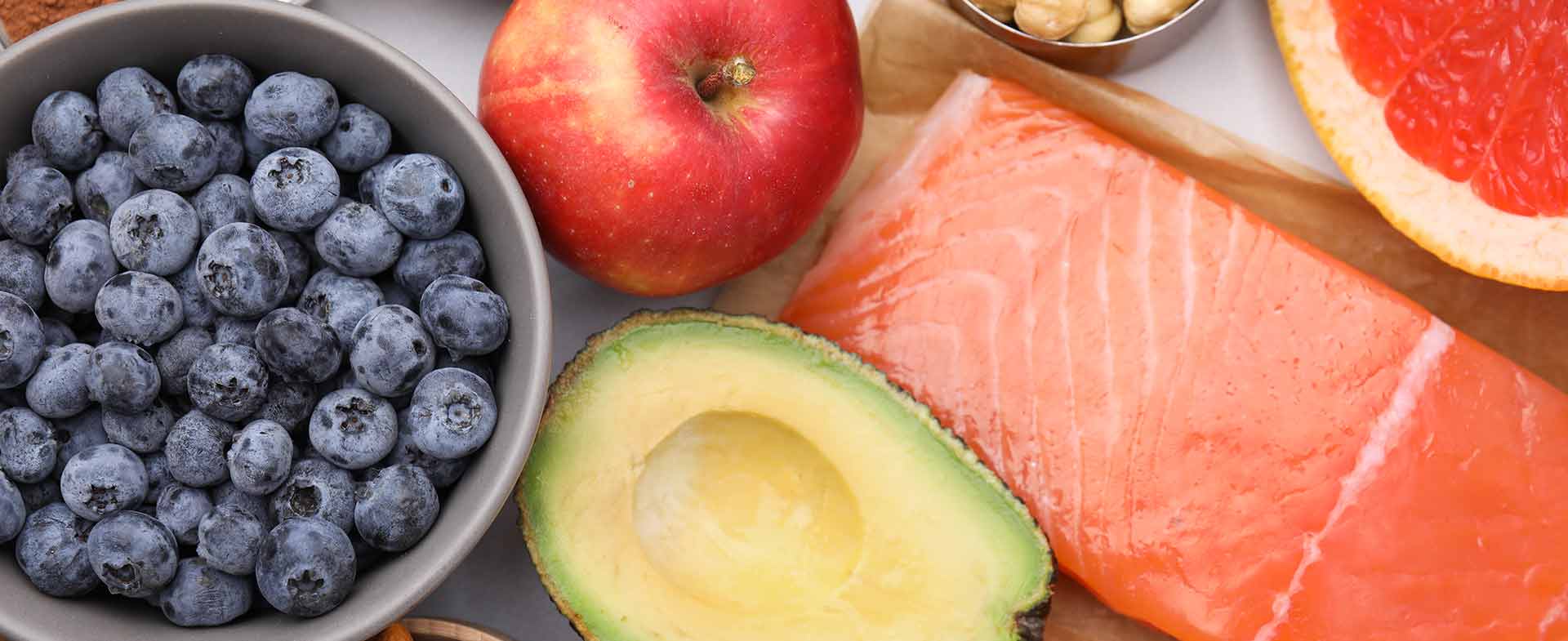A variety of nutritious foods are displayed, including a raw salmon fillet, avocado, blueberries, apple, kiwi, grapefruit half, almonds, hazelnuts, cocoa powder, and a spoonful of red caviar. meal timing for heart health