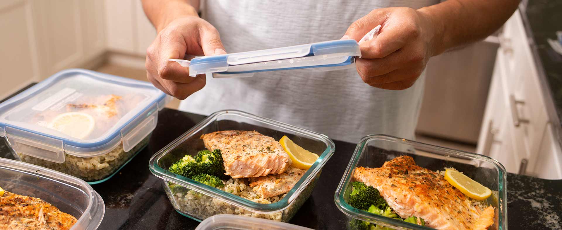 A person in a kitchen is sealing a food container filled with broccoli and salmon. Additional containers with similar meals are on the counter. new dietary guidelines