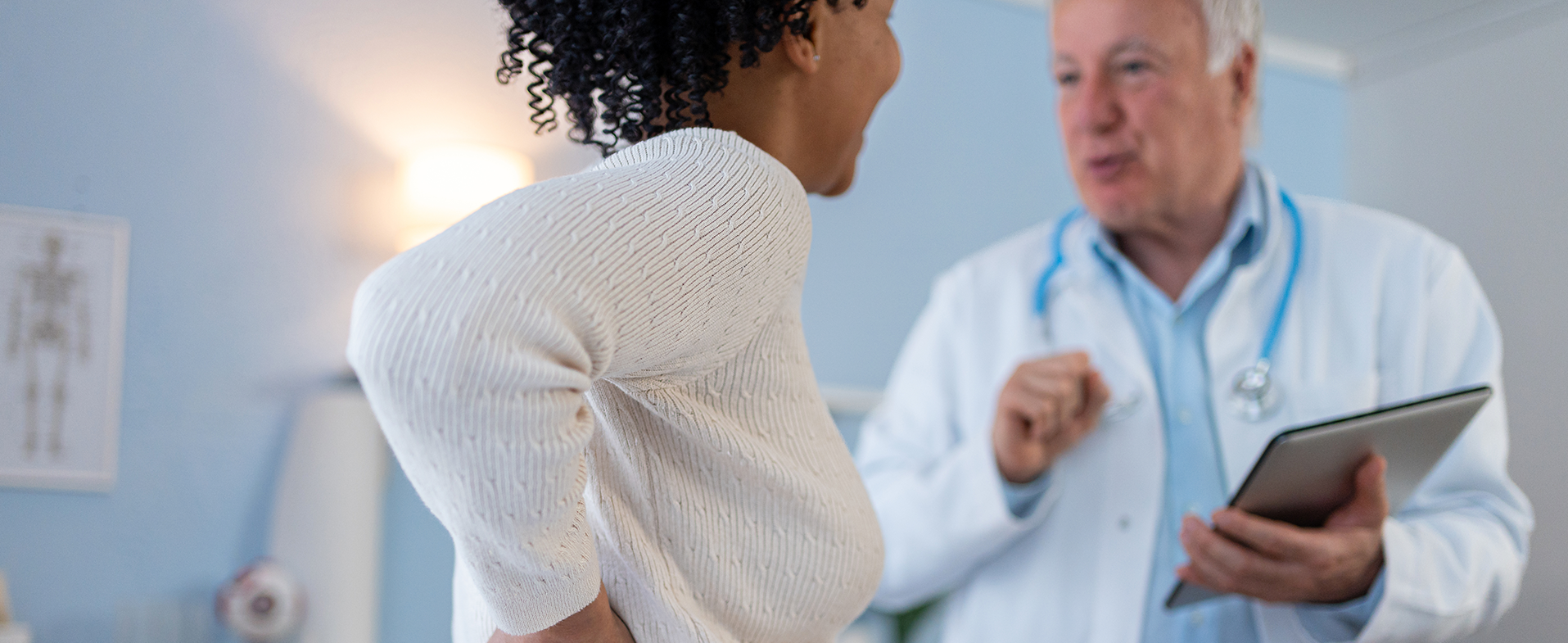 Middle-aged woman wearing a long-sleeved cream sweater and holding her back. She is sitting down talking to a male doctor who is wearing a white coat and a blue button-down shirt. He is holding a clipboard. dummy desktop Image