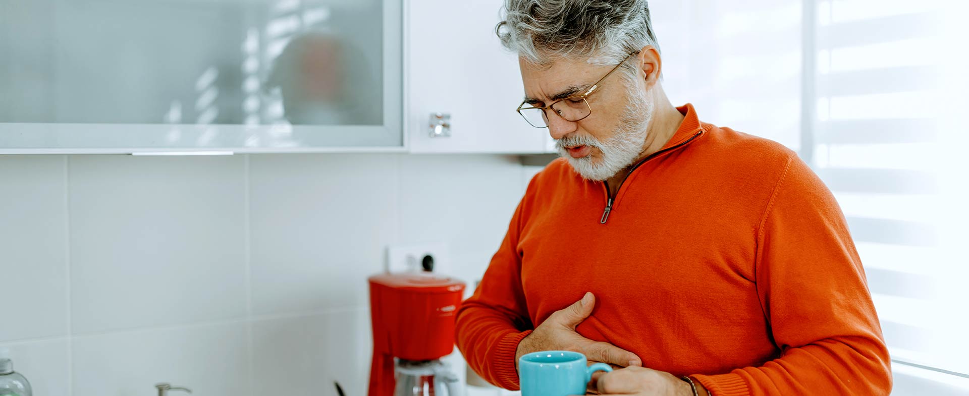 A man with a gray beard wearing an orange sweater stands in a kitchen holding a blue mug. He appears to be experiencing discomfort, with one hand on their chest. A red coffee maker and various kitchen items are on the countertop nearby. gerd