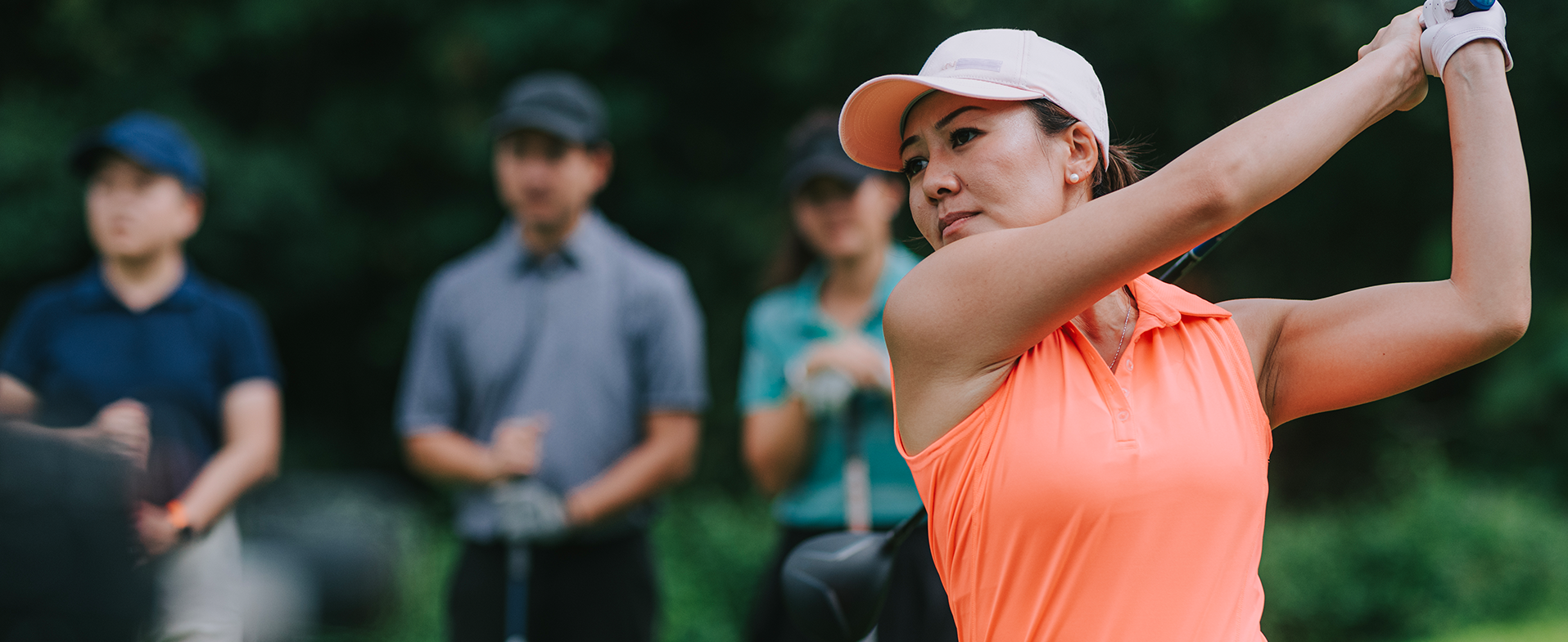 Female athlete outside playing golf. She is wearing a white visor on her head and a neon orange sleeveless, collared athletic shirt. Two golfers - a male and female - are standing in the background watching her. dummy desktop Image