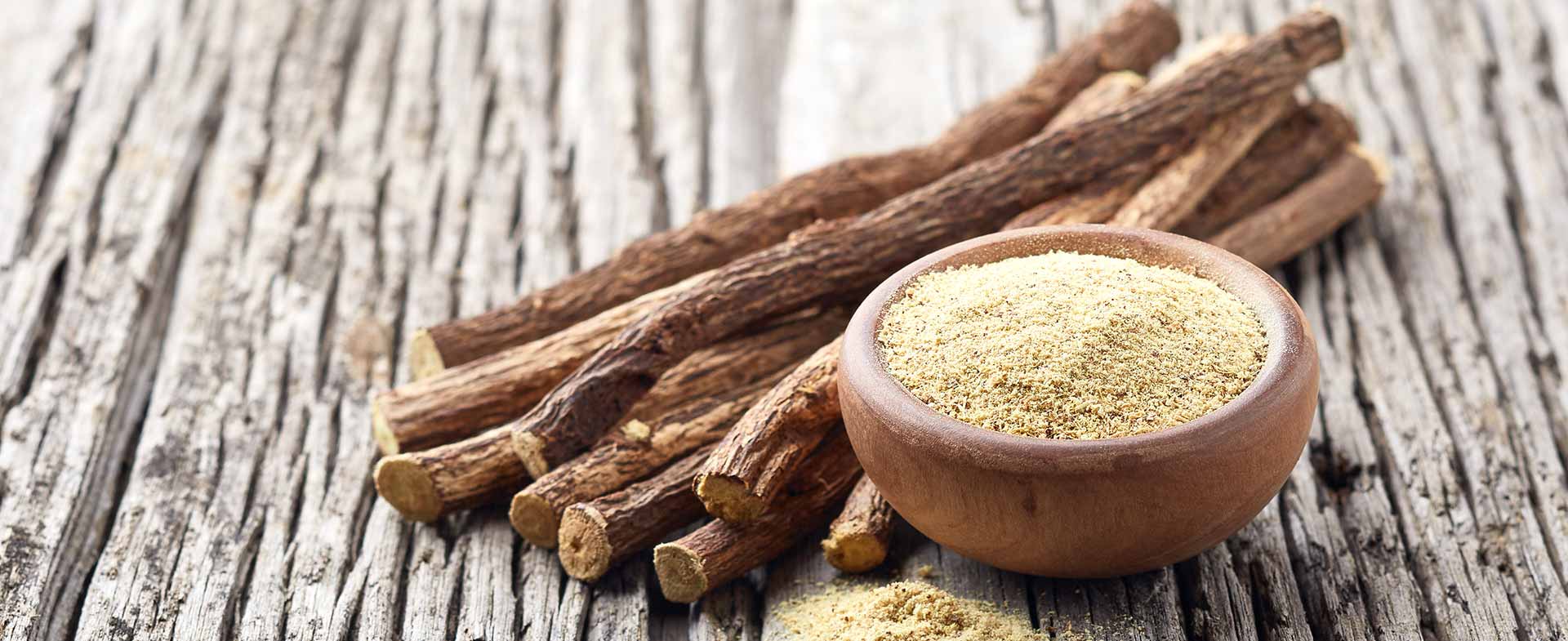 A small wooden bowl filled with ground licorice powder is placed on a rustic wooden surface. Several whole licorice roots are arranged beside the bowl. licorice root and heart health