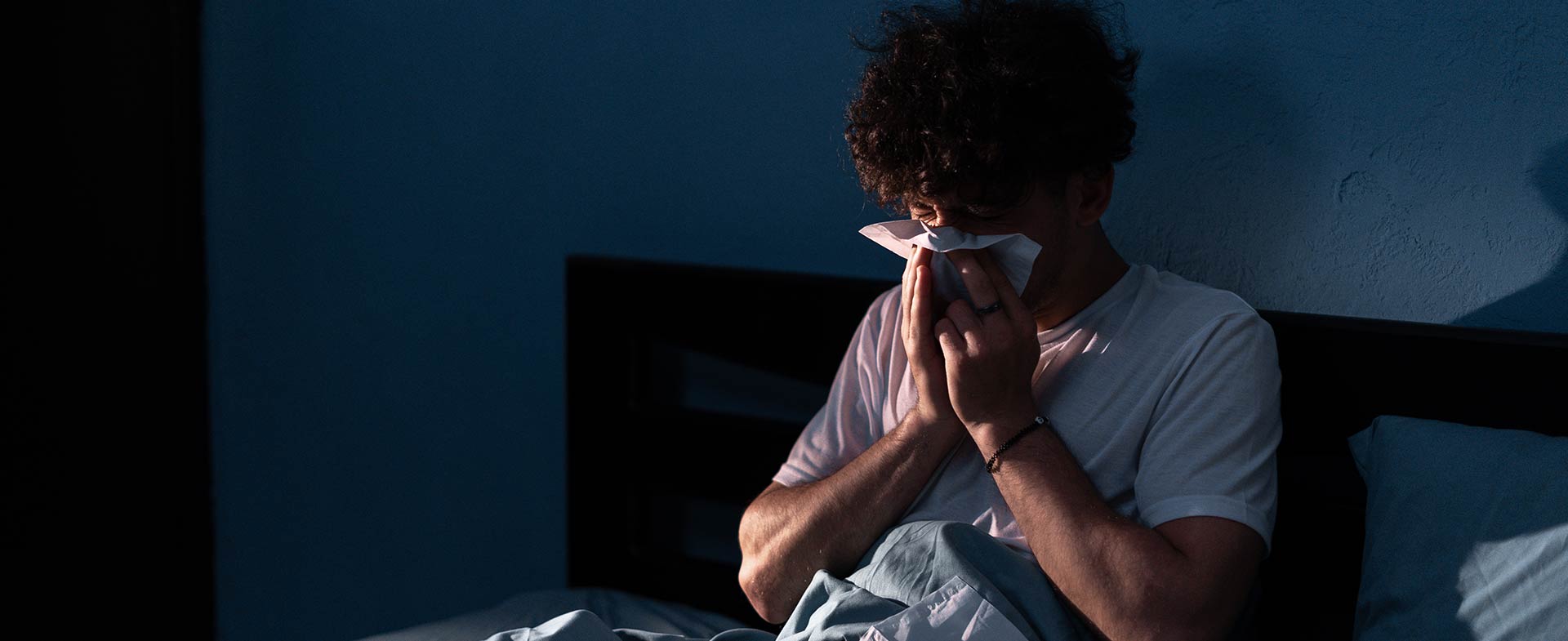 A person sitting up in bed in a dimly lit room, using a tissue to blow their nose. The bedding is a muted blue color. nighttime congestion