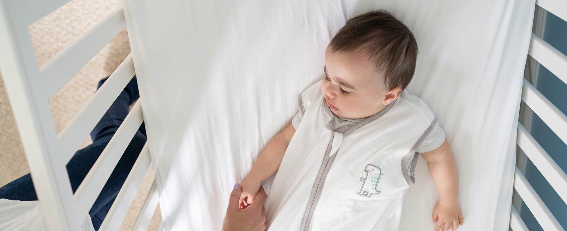 A baby lying in a crib, wearing a white sleep sack with a small dinosaur design. An adult hand is gently touching the baby's hand. sound machines infant sleep
