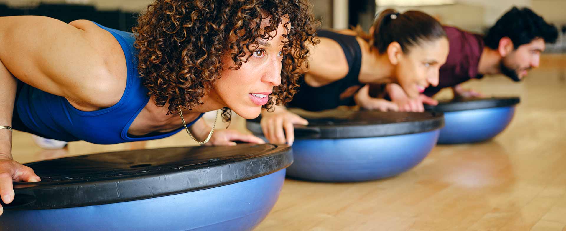 Three individuals are exercising on Bosu balls in a gym, focusing intently on holding a pushup position. The room has wooden floors and large mirrors. workout gadgets