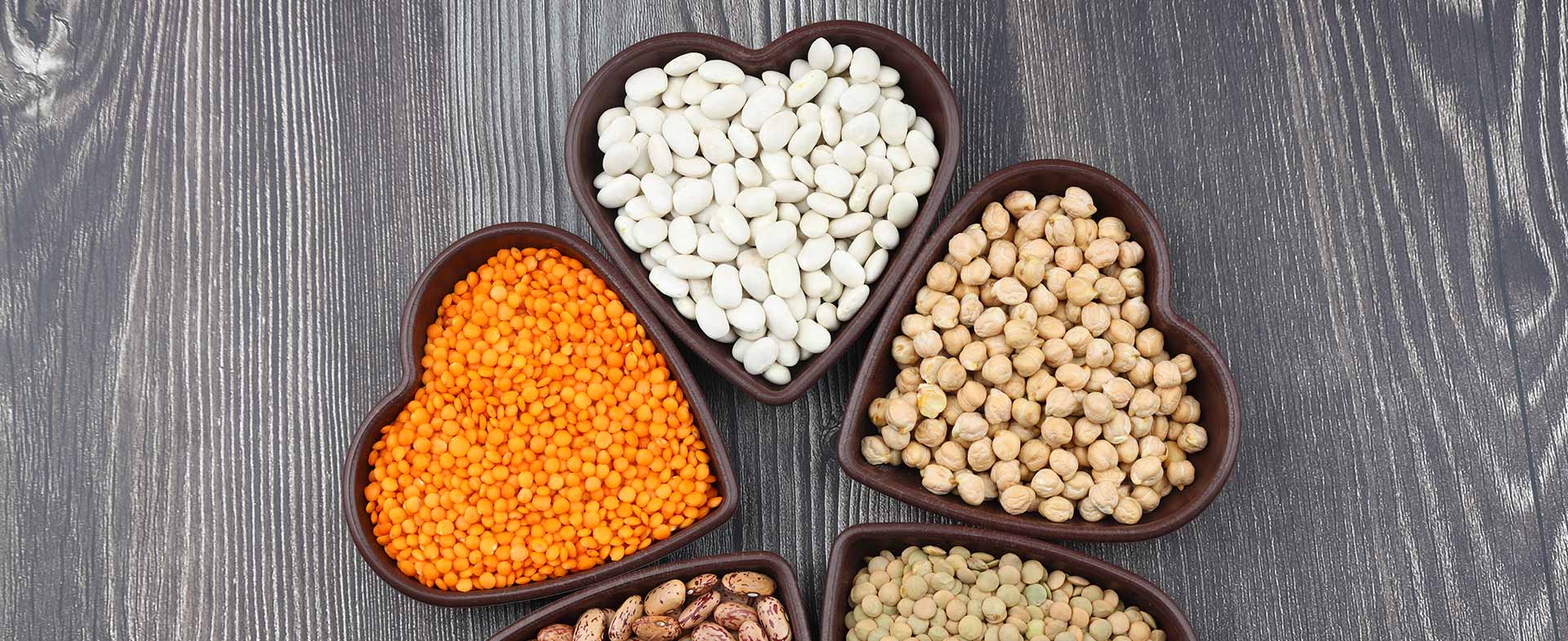 Heart-shaped bowls contain a variety of legumes on a dark wooden surface. The bowls hold white beans, red lentils, chickpeas, pinto beans, and green lentils. beans for blood pressure