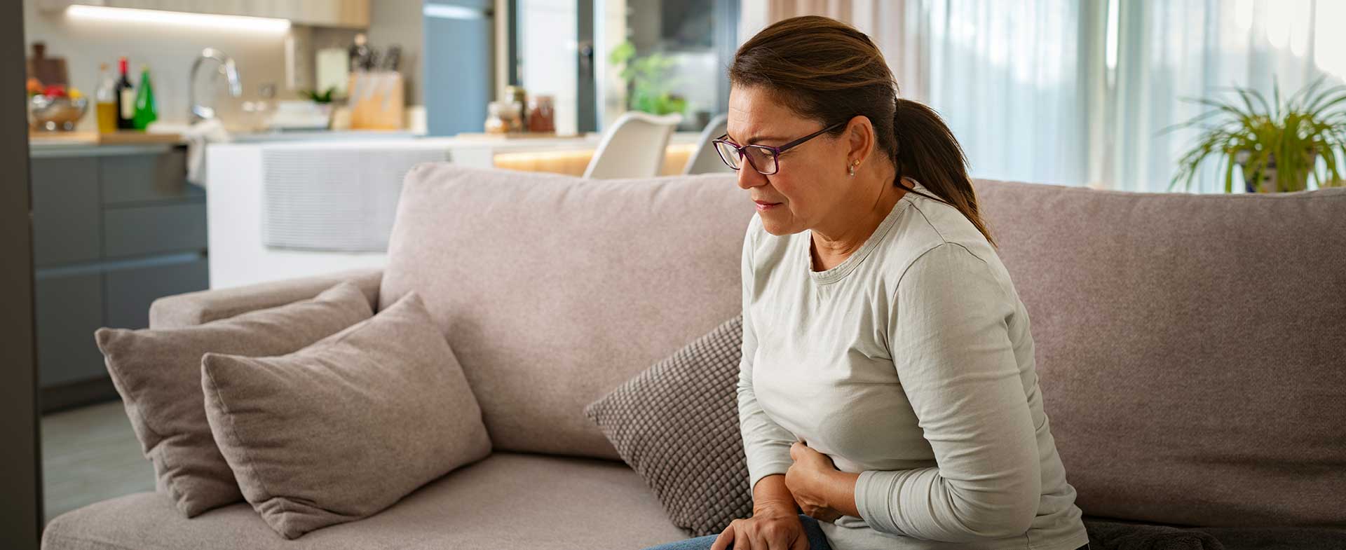 A person sitting on a sofa in a modern living room, holding their stomach and appearing uncomfortable. cyclosporiasis outbreak