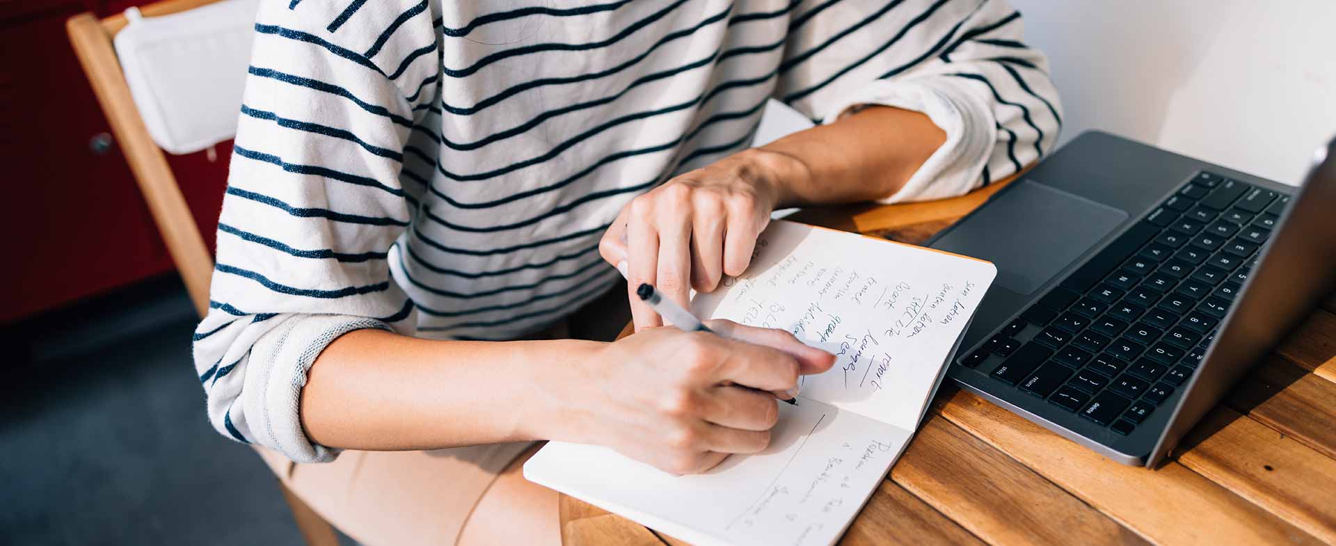 A person in a striped shirt writes notes in a notebook while sitting at a wooden table with a laptop open. handwriting vs typing