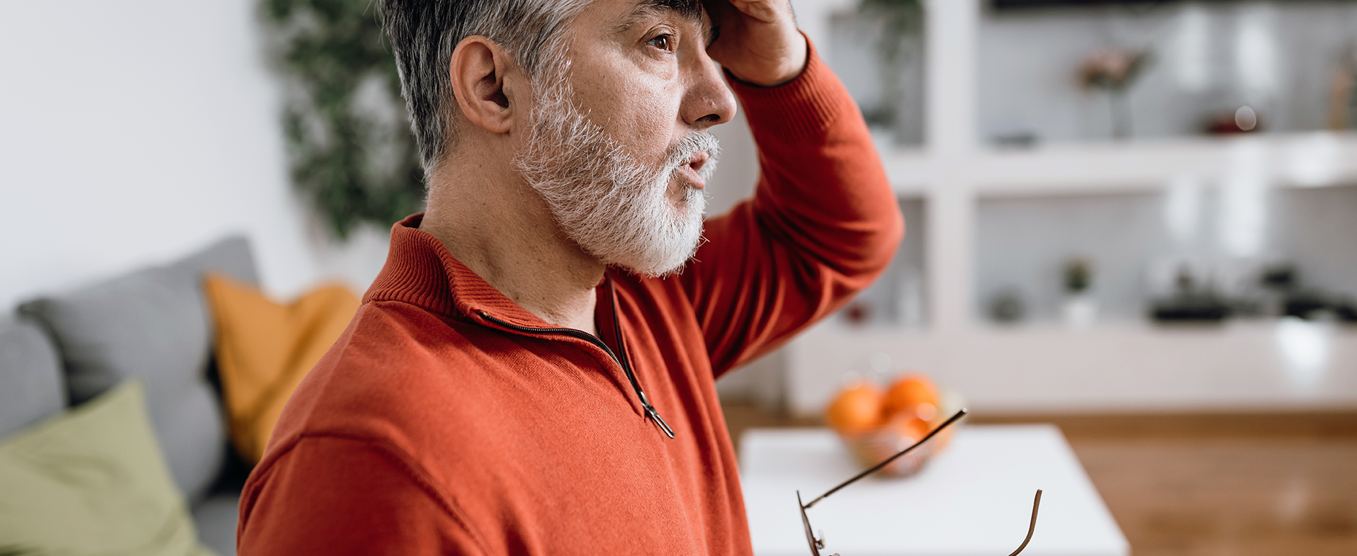 Man wearing orange half-zip sweater with a gray beard and gray hair. He has his hand on his forehead and holding his glasses in his other hand. dummy desktop Image