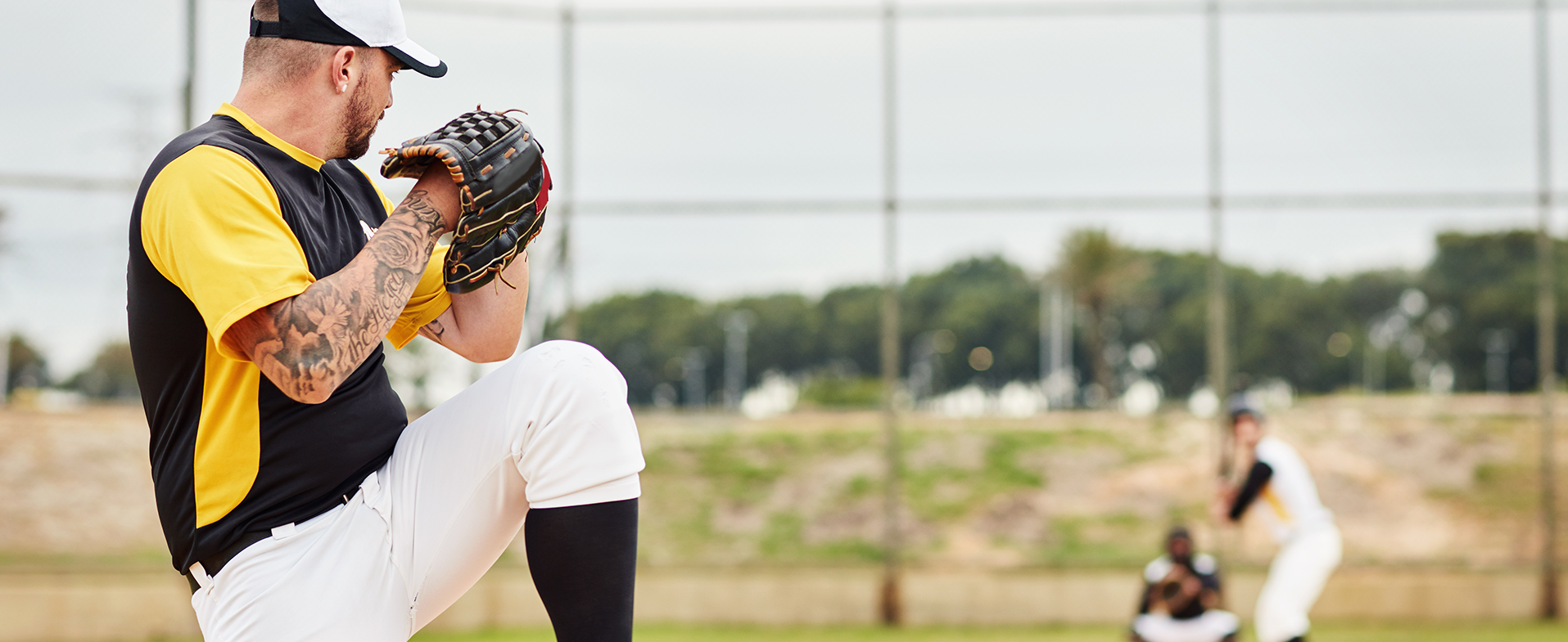 Baseball pitcher on mound winding up a pitch. He is wearing a black and white baseball hat, a black and white t-shirt and white baseball pants. He has an arm sleeve of tattoos. dummy desktop Image