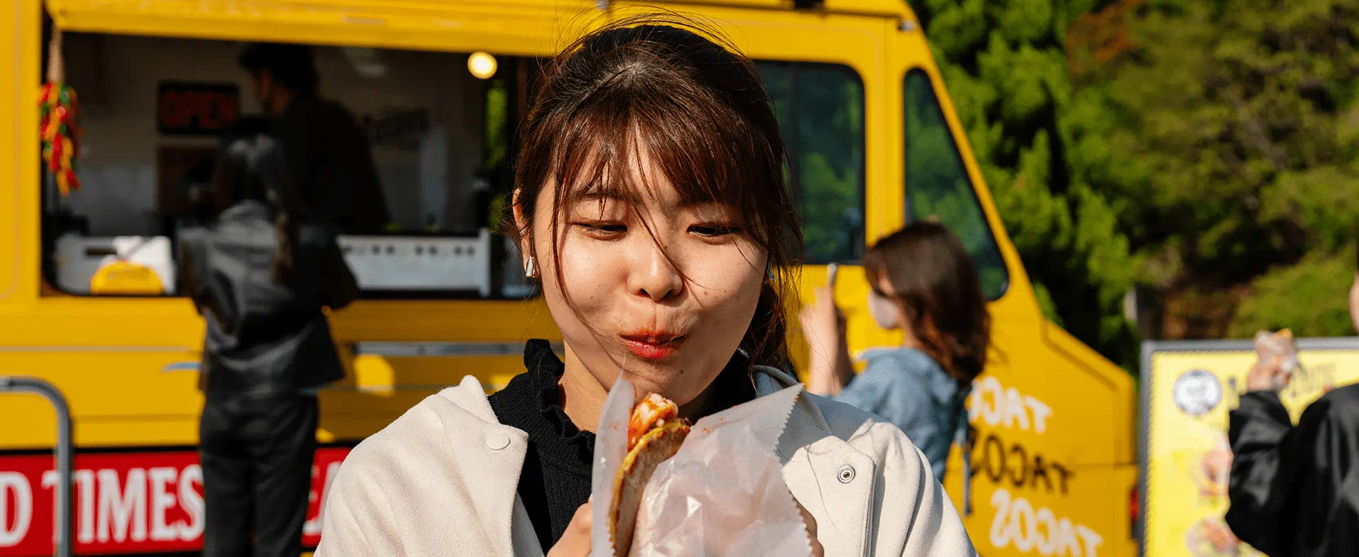 Young woman with dark hair and bangs standing outside in front of a yellow taco truck eating a taco. woman eating taco
