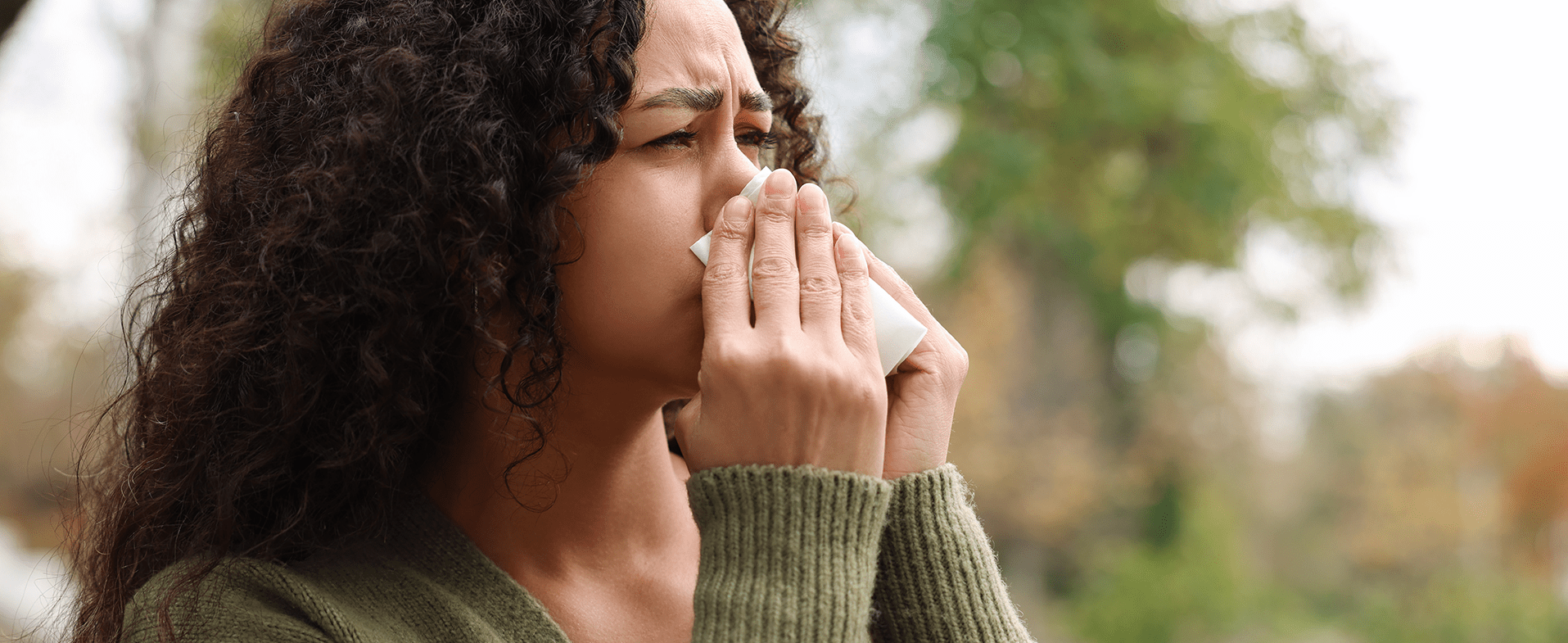 Woman with dark curly hair wearing a green long-sleeve sweater and blowing her nose outside. dummy desktop Image