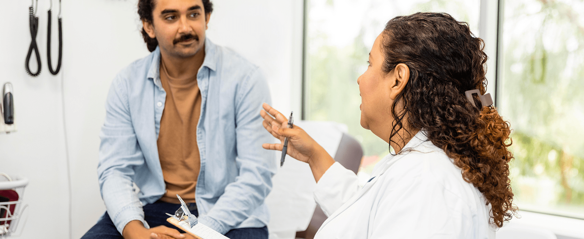 Man with dark hair and a moustache sitting in a doctor's office. He is wearing a light blue button-down shirt with a tan t-shirt underneath, listening to a female doctor talk to him. She has brown curly hair and is wearing a lab coat. dummy desktop Image