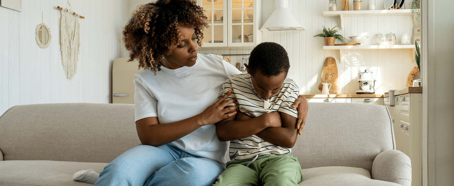 Mom with brown curly short hair wearing a white T-shirt and jeans. She is sitting on a couch trying to comfort her young son who is wearing a white and navy blue striped t-shirt and green pants. He has his arms folded over his chest. dummy desktop Image