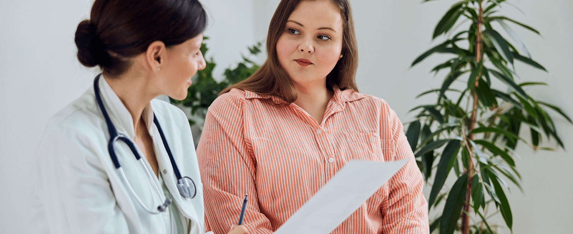 Female doctor with dark brown hair in a low bun wearing a white coat and a stethoscope around her neck. She is talking to a young female patient with long brown hair who is wearing an orange and white vertical striped button-down shirt. dummy desktop Image