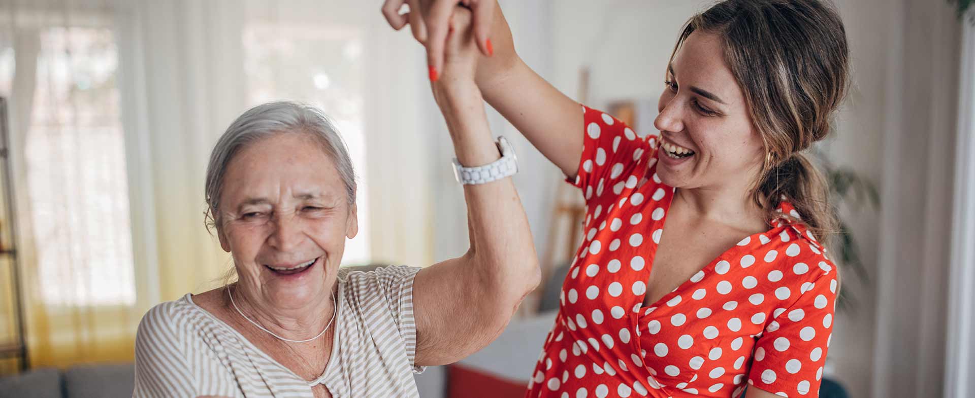 Two women are smiling and dancing indoors. One person is older with grey hair and the other is younger wearing a red polka dot top. The setting appears to be a bright room with curtains in the background. everyday exercises for brain health