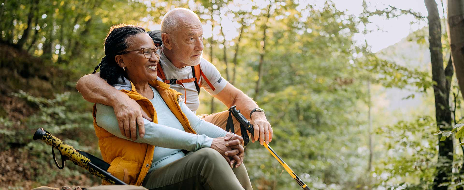 A man with short white hair and a backpack is sitting on a hillside, arm around a woman with long braided hair and glasses. Both are smiling and holding hiking poles, surrounded by lush green trees and sunlight. preventing osteoporosis