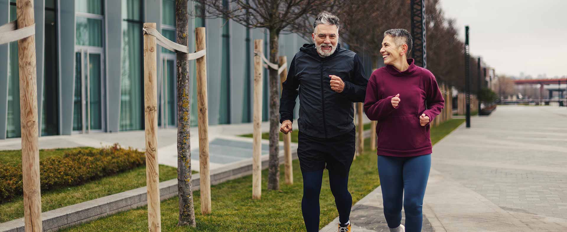 Two people jogging on a sidewalk next to a modern building. They appear to be enjoying the activity together. Trees are planted along the path. The weather is overcast. walking speed cognitive health