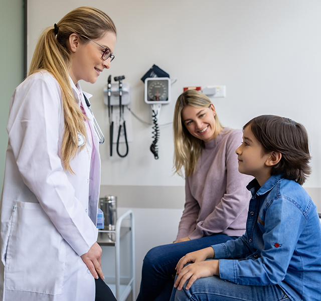 Little boy wearing blue button down shirt at pediatrician appointment with his mom who has long blonde hair and is wearing a lilac sweater. The pediatrician is wearing a white coat with glasses and has her hair in a ponytail. dummy mobile image