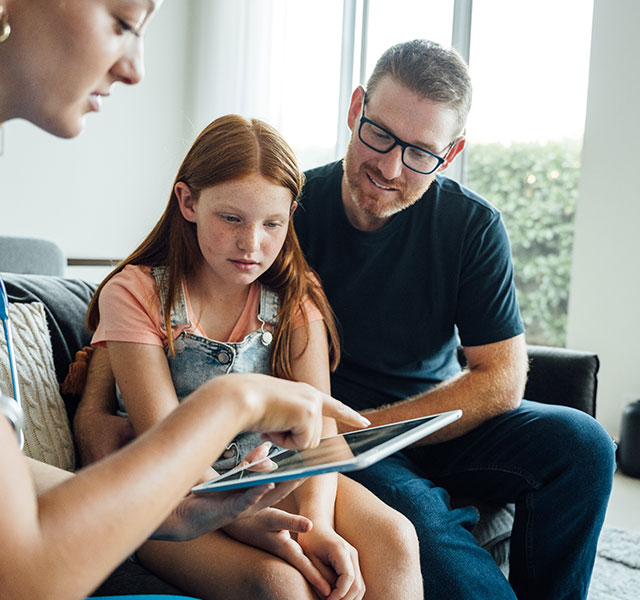 A father with facial hair and glasses sits on a couch with his pre-teen redhead daughter. They are sitting with a nurse in a blue top who is reviewing information with them on a tablet. autism healthcare transition
