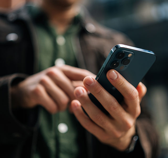 A person is holding and using an iPhone outdoors, wearing a dark jacket and a green shirt. The background is slightly blurred. dystextia