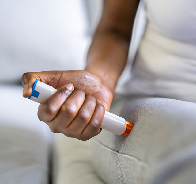 A person seated on a couch is using an epinephrine auto-injector (EpiPen) on their thigh, depicted in a close-up shot. The individual wears a light-colored shirt and pants. epipen interactions