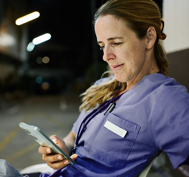 A woman with a blonde ponytail in medical scrubs is seated outdoors, looking at a smartphone. The background suggests a hospital environment at night. digital interventions for night shift workers