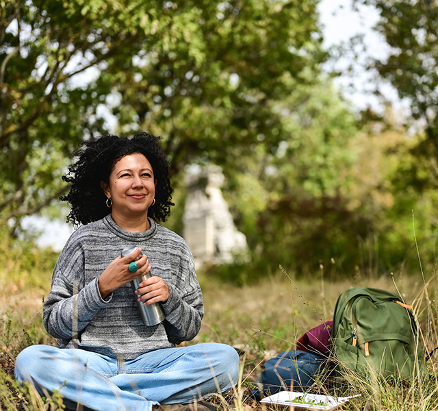 Woman with dark shoulder-length curly hair sitting outside in the grass cross-legged. She is holding a metal water bottle and wearing a gray striped sweater and jeans. dummy mobile image