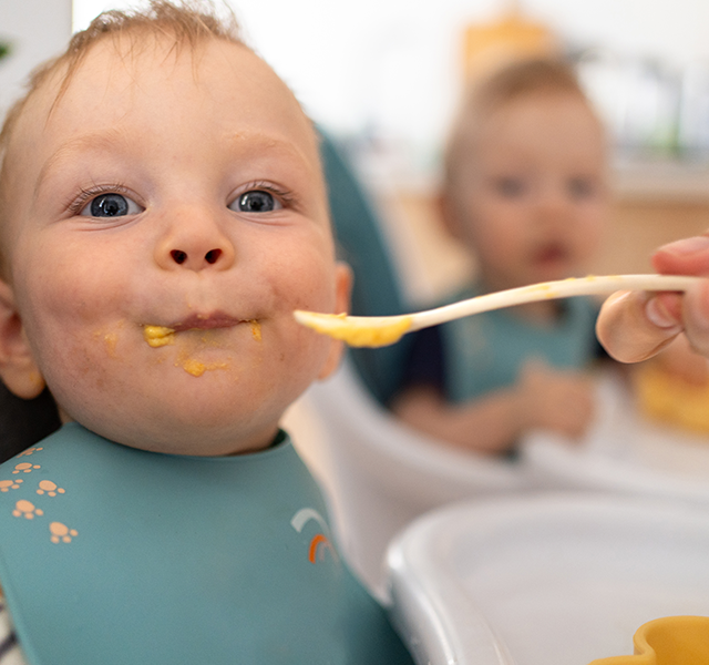 Baby eating an orange-ish yellow colored puree from a spoon and wearing a blue bib. An adult hand is holding the spoon. dummy mobile image