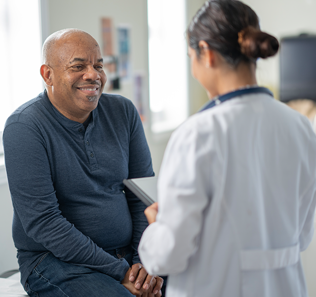 Bald man wearing navy long-sleeved henley shirt smiling and talking to doctor. Her hair is in a low bun and she is wearing a white coat. doctor talking to patient