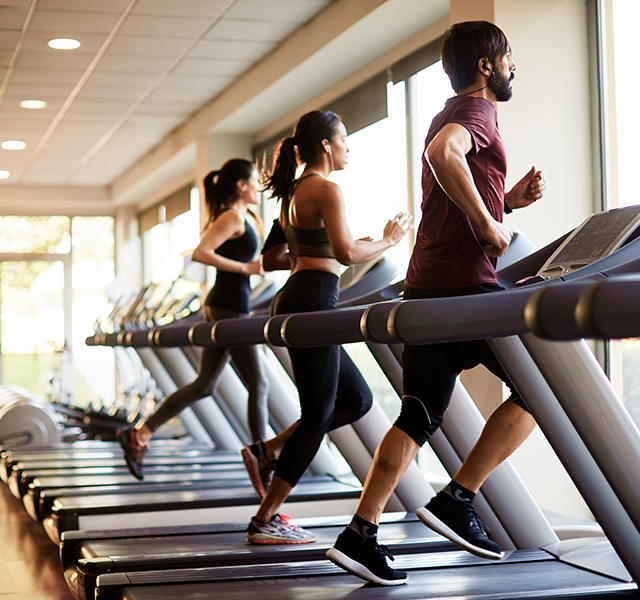 Two women and one male at a gym running on treadmills. They are looking out the window and wearing workout clothes. people running on treadmill