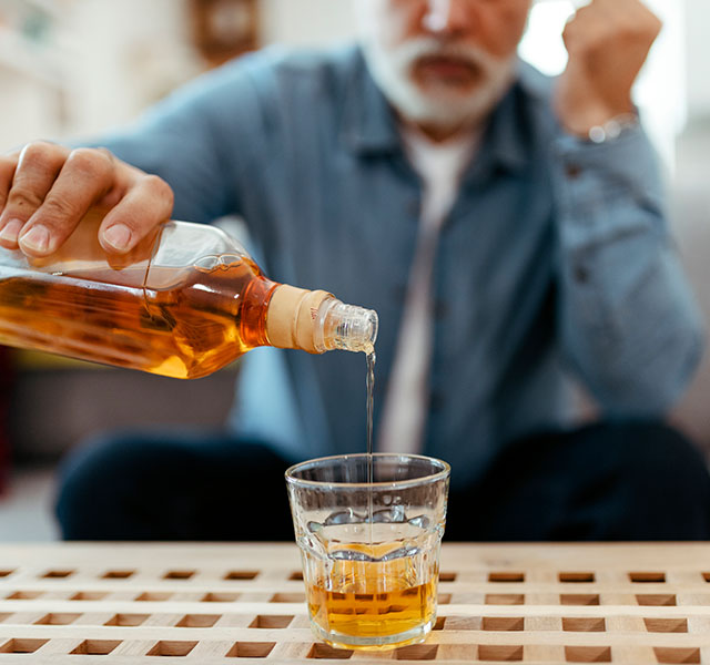A man with a white beard pours a dark liquor such as whiskey or rum from a bottle into a glass on a wooden table. The background shows a sofa with cushions. heavy drinking and stroke