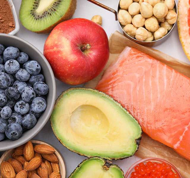 A variety of nutritious foods are displayed, including a raw salmon fillet, avocado, blueberries, apple, kiwi, grapefruit half, almonds, hazelnuts, cocoa powder, and a spoonful of red caviar. meal timing for heart health