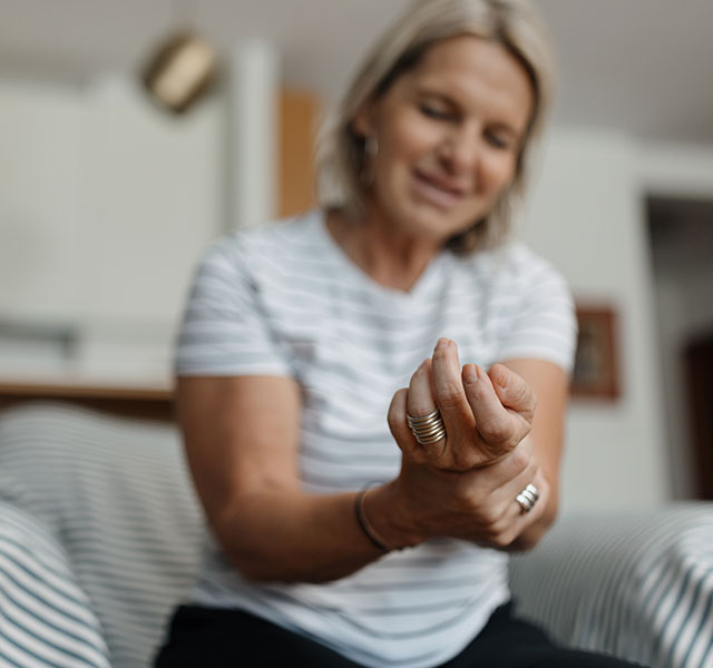 A blond woman in a striped short-sleeve shirt sits while holding her wrist in discomfort. muscles spasms after stroke