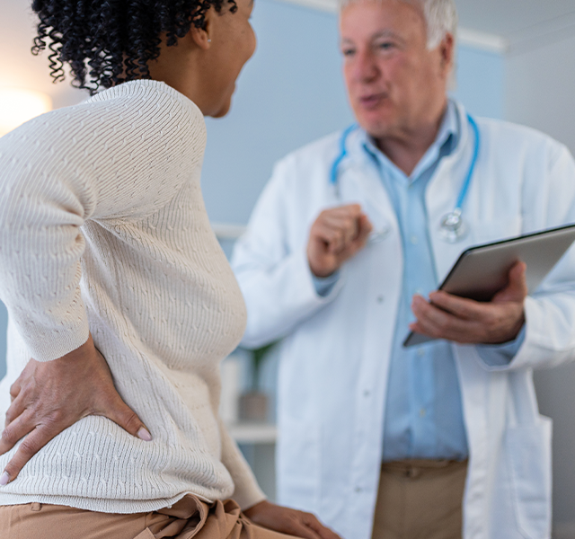 Middle-aged woman wearing a long-sleeved cream sweater and holding her back. She is sitting down talking to a male doctor who is wearing a white coat and a blue button-down shirt. He is holding a clipboard. dummy mobile image