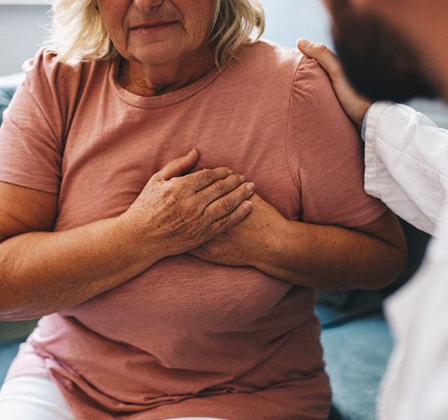 A woman in a pink shirt is seated on a couch with her hands over her heart. A male doctor sits opposite of her with a hand on her shoulder in comfort. pain after heart attack