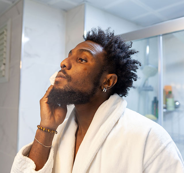 A man with dark skin in a white bathrobe is standing in a bathroom, cleansing their face. The background shows a tiled wall and a glass shower door. skin conditions in dark skin