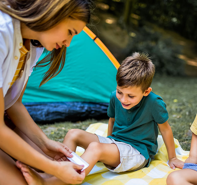 A young woman tends to a child's scraped knee. They are seated with another child on a picnic blanket near a green and orange tent in a grassy area. summer camp ailments