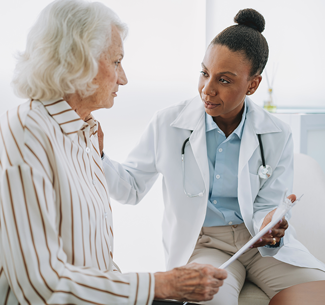 Older woman with white hair and white button-down shirt with vertical brown stripes talking to female doctor wearing a white coat, blue button-down shirt and hair in a bun. doctor talking to patient