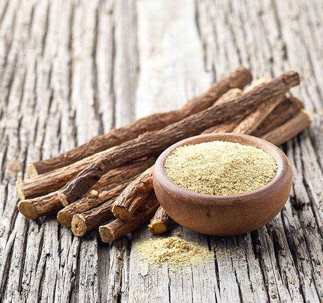 A small wooden bowl filled with ground licorice powder is placed on a rustic wooden surface. Several whole licorice roots are arranged beside the bowl. licorice root and heart health