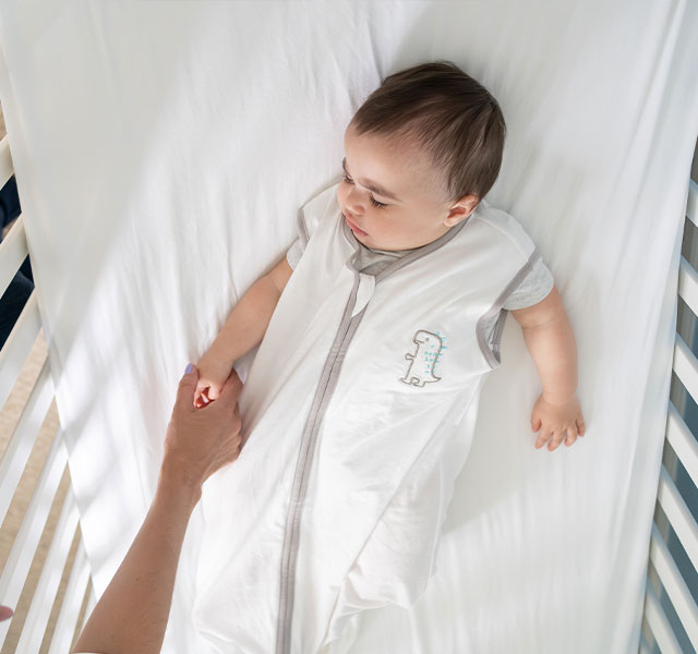 A baby lying in a crib, wearing a white sleep sack with a small dinosaur design. An adult hand is gently touching the baby's hand. sound machines infant sleep