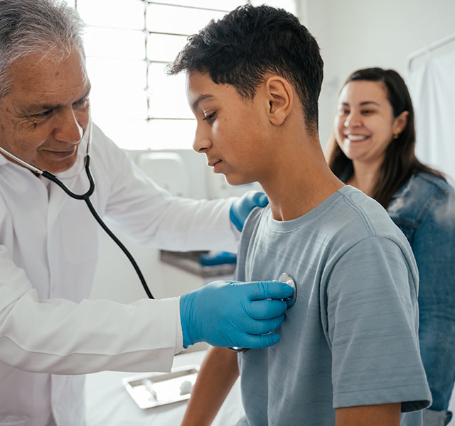 A medical professional uses a stethoscope to listen to a patient's chest during a check-up. Another person is smiling in the background. The setting appears to be a clinic or doctor's office. baseline heart testing for student athletes